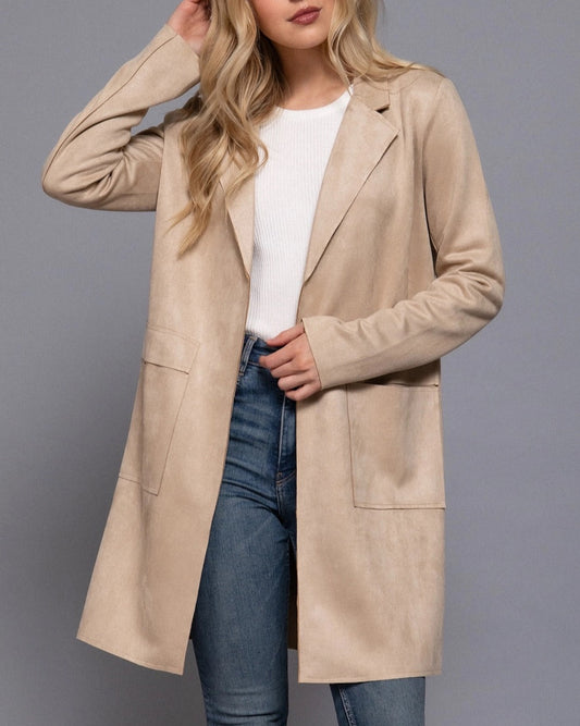 Woman wearing a sand faux suede coat over a white shirt and blue jeans on a gray background.