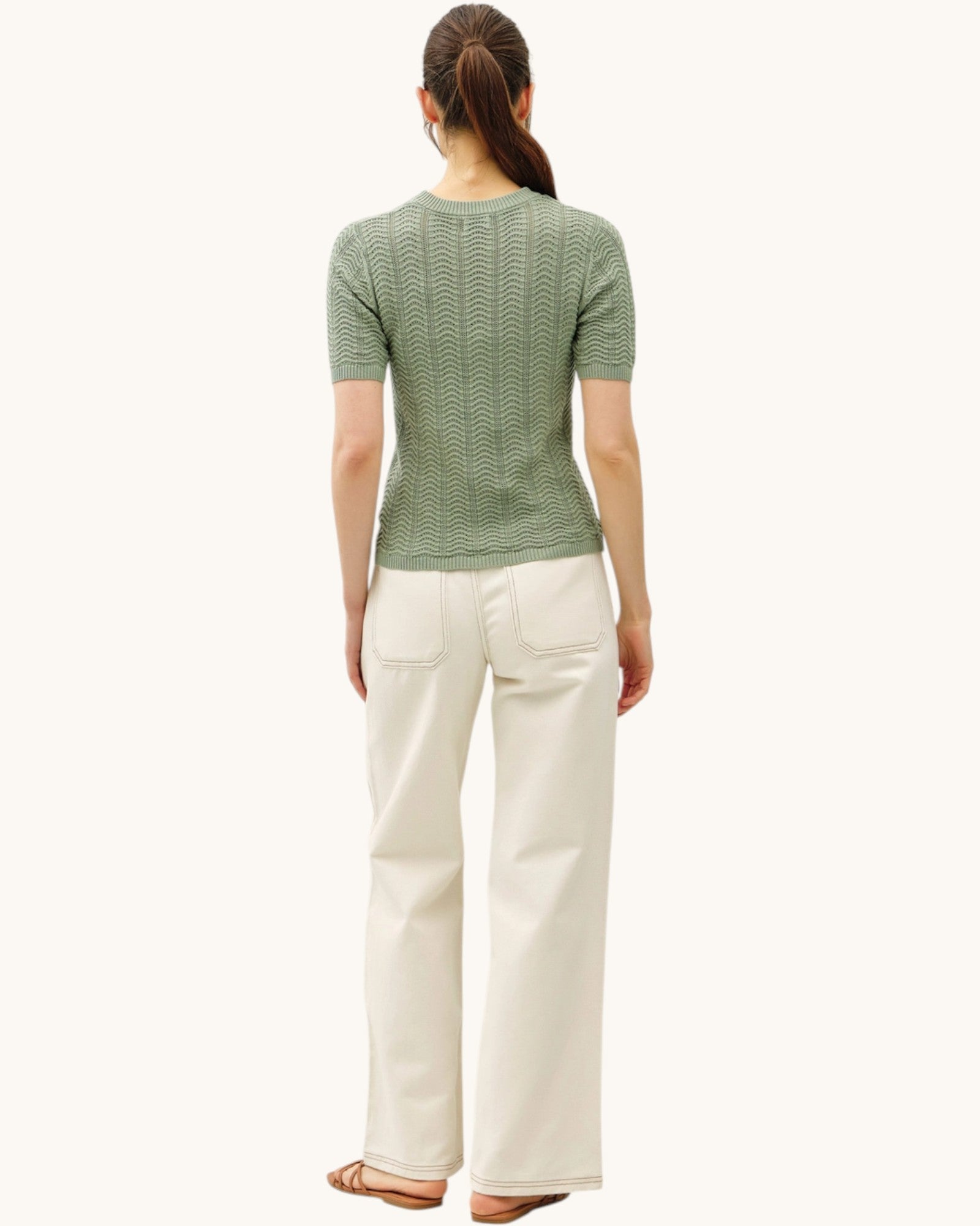 Back  view of a woman wearing a green patterned chevron short sleeve sweater and khaki pants on a white background.