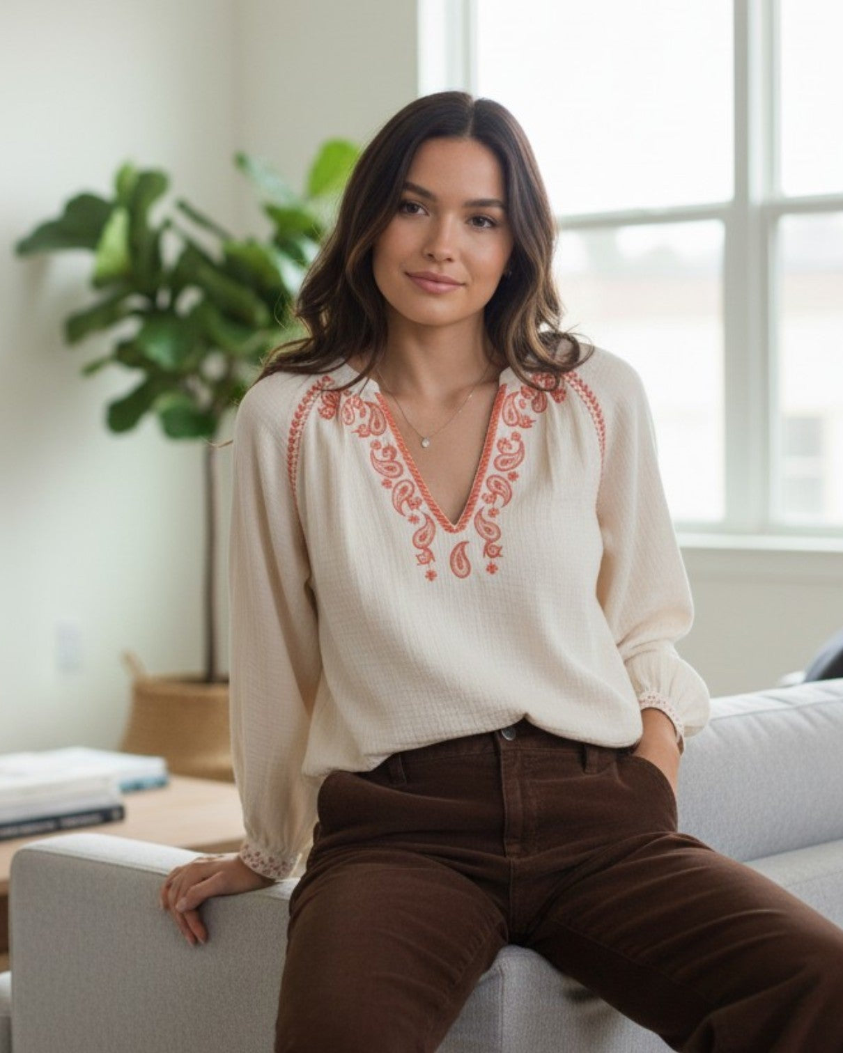 A woman sits on a light gray sofa in a bright living room, wearing a cream long-sleeve blouse with coral-orange embroidered detailing around the V-neck and brown corduroy pants.
