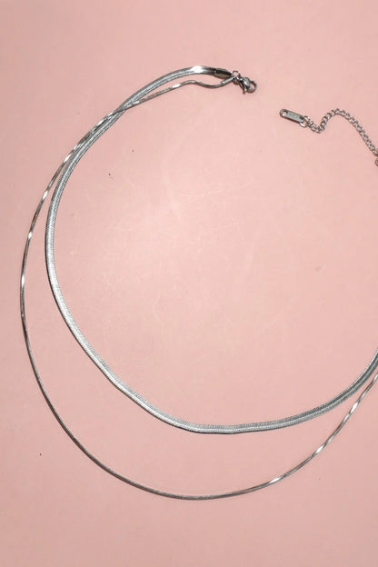 A silver-toned snake and herringbone double layer chain necklace with a lobster clasp and extender chain, displayed on a solid salmon pink background.