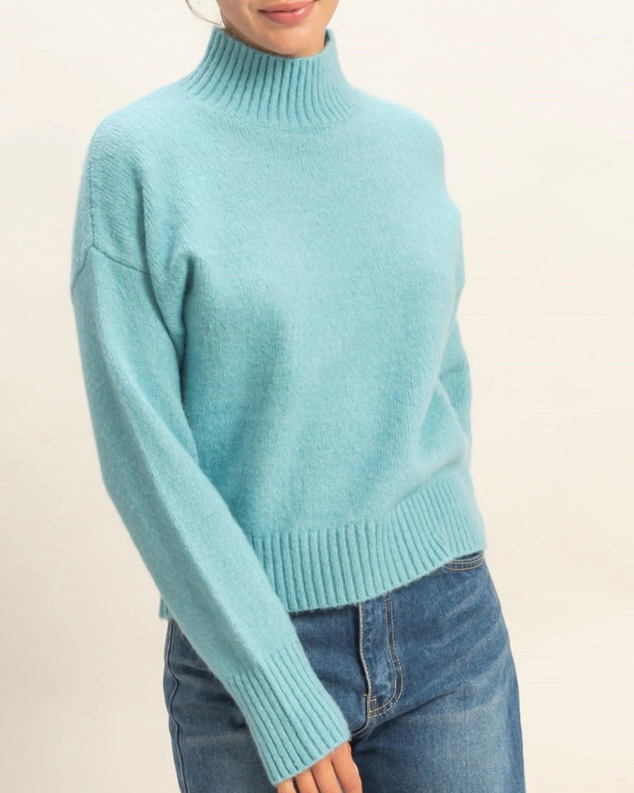 Person wearing a sky blue turtleneck sweater and jeans on a beige background.