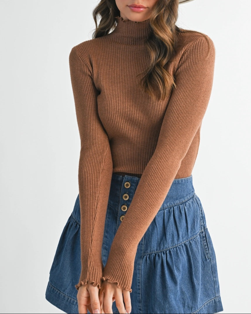 Woman wearing a brown ribbed long-sleeve top and blue denim skirt with a white hat on a plain background
