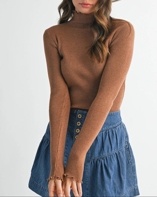 Woman wearing a brown ribbed long-sleeve top and blue denim skirt with a white hat on a plain background