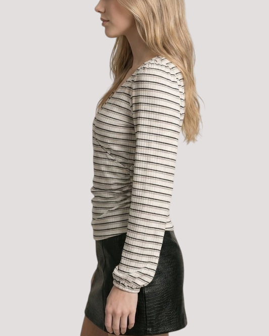 A woman shown in side profile wearing a fitted long-sleeve ribbed top with thin horizontal stripes in beige and black paired with a black leather mini skirt, against a light gray studio background.
