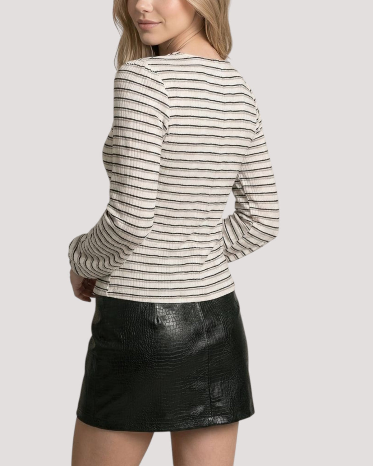 A woman shown from the back wearing a fitted ivory long-sleeve top with thin horizontal black and beige stripes paired with a black textured leather mini skirt, against a light gray studio background.