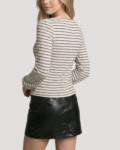 A woman shown from the back wearing a fitted ivory long-sleeve top with thin horizontal black and beige stripes paired with a black textured leather mini skirt, against a light gray studio background.