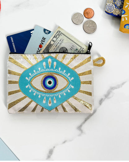 Small zipper pouch/coin purse with eye design with sunburst pattern, money, and coins on a marble surface.