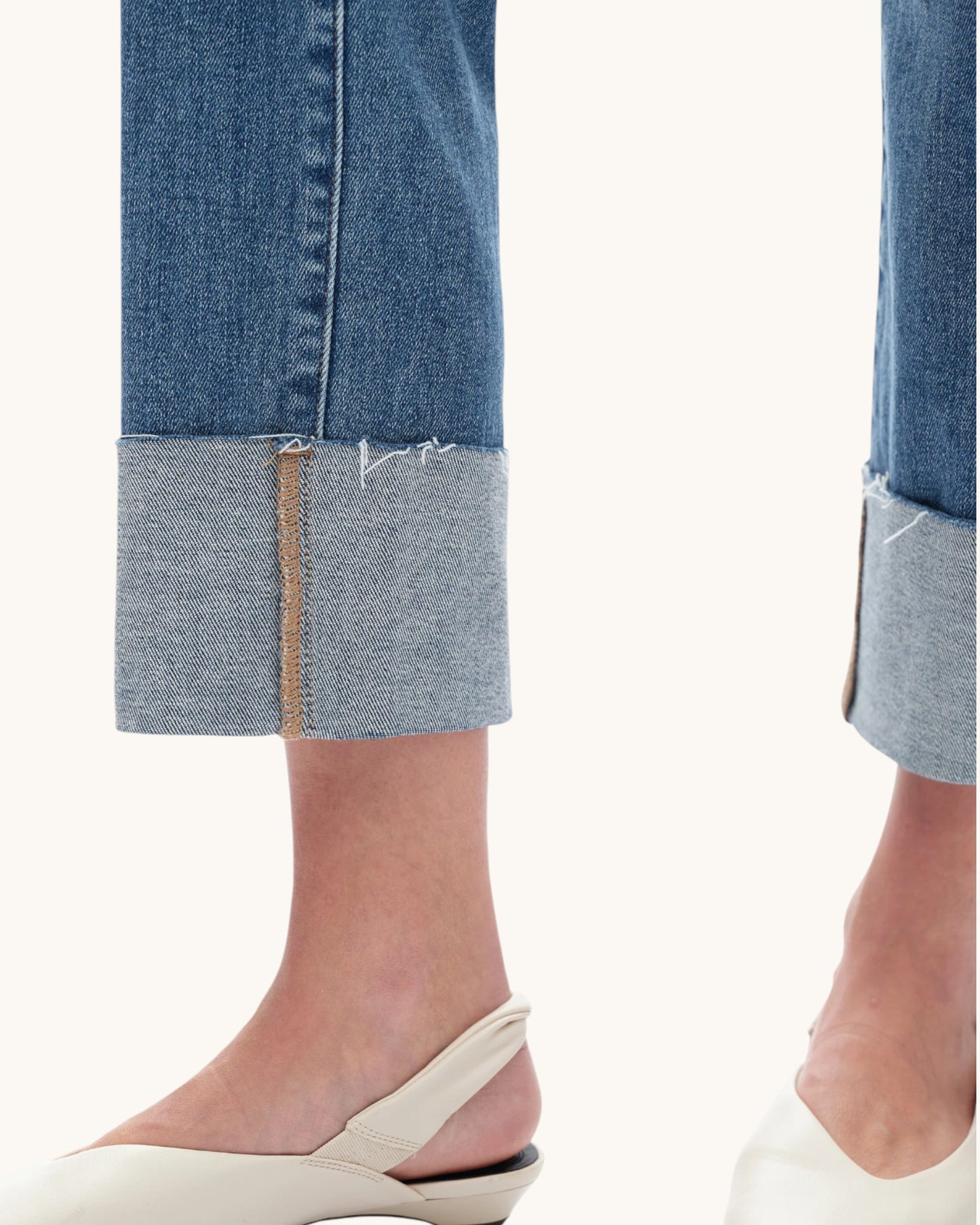Close-up of a person's lower legs wearing blue denim jeans with rolled cuffs and off-white slingback flat shoes against a plain light background.