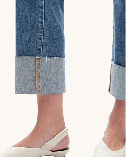 Close-up of a person's lower legs wearing blue denim jeans with rolled cuffs and off-white slingback flat shoes against a plain light background.