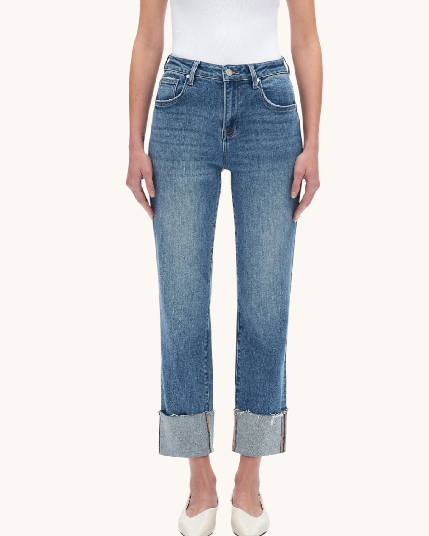 A model wearing a pair of medium-wash blue cropped jeans with rolled cuffs, paired with a white top and white pointed-toe heels, against a solid off-white background.