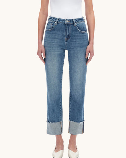 A model wearing a pair of medium-wash blue cropped jeans with rolled cuffs, paired with a white top and white pointed-toe heels, against a solid off-white background.