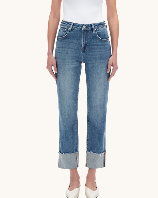 A model wearing a pair of medium-wash blue cropped jeans with rolled cuffs, paired with a white top and white pointed-toe heels, against a solid off-white background.