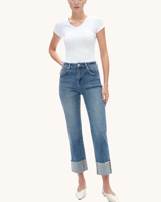 A model wearing a white short-sleeve V-neck t-shirt and high-waisted blue cropped jeans with rolled cuffs, paired with white pointed-toe heels, against a solid off-white background.
