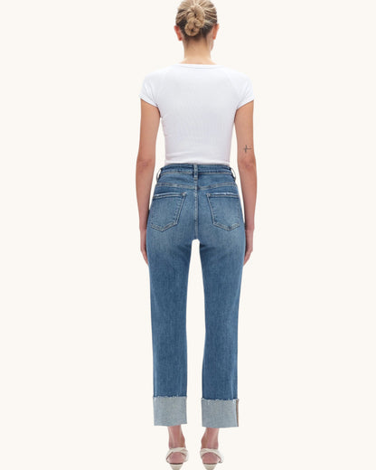 A woman viewed from the back wearing a white short-sleeve t-shirt and high-waisted blue jeans with rolled cuffs, set against a solid off-white background.
