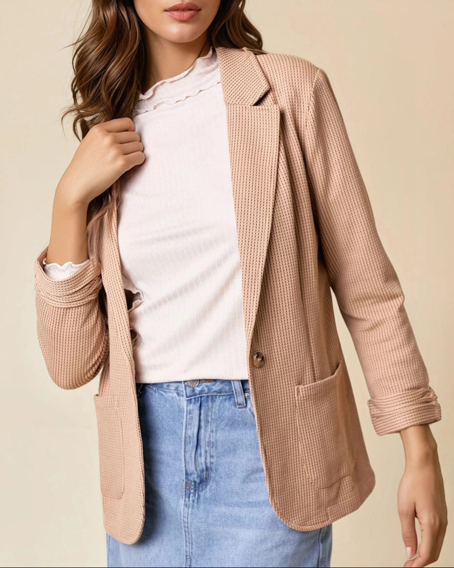 Woman wearing a tan blazer over a white shirt and blue jeans on a beige background.