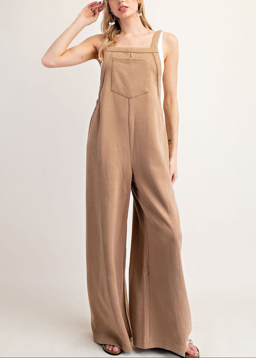 A woman modeling a sleeveless, wide-leg jumpsuit in a warm taupe-brown color with a subtle textured pattern, set against a plain light gray studio background.