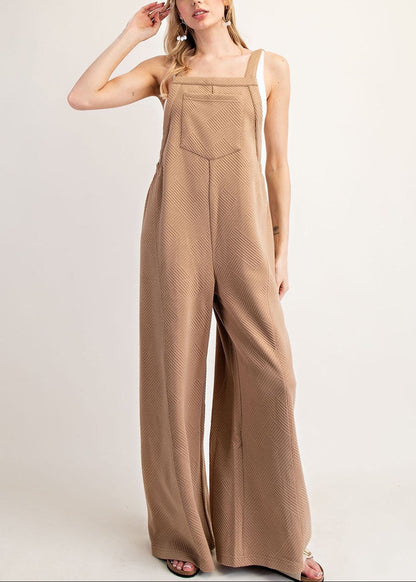 A woman modeling a sleeveless, wide-leg jumpsuit in a warm taupe-brown color with a subtle textured pattern, set against a plain light gray studio background.