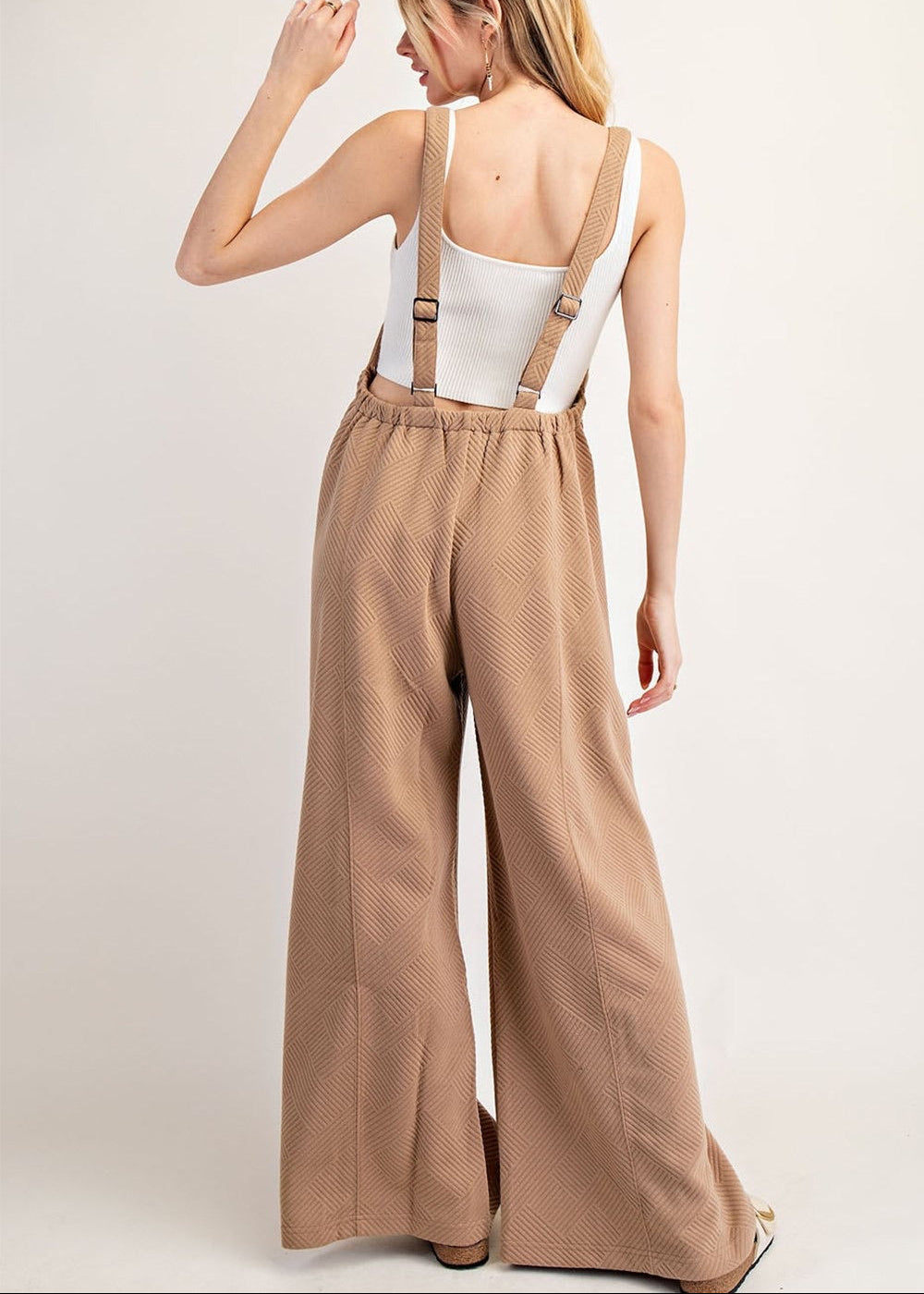 A woman shown from the back wearing a wide-leg tan jumpsuit with adjustable suspenders over a white tank top, photographed against a plain light beige studio background.