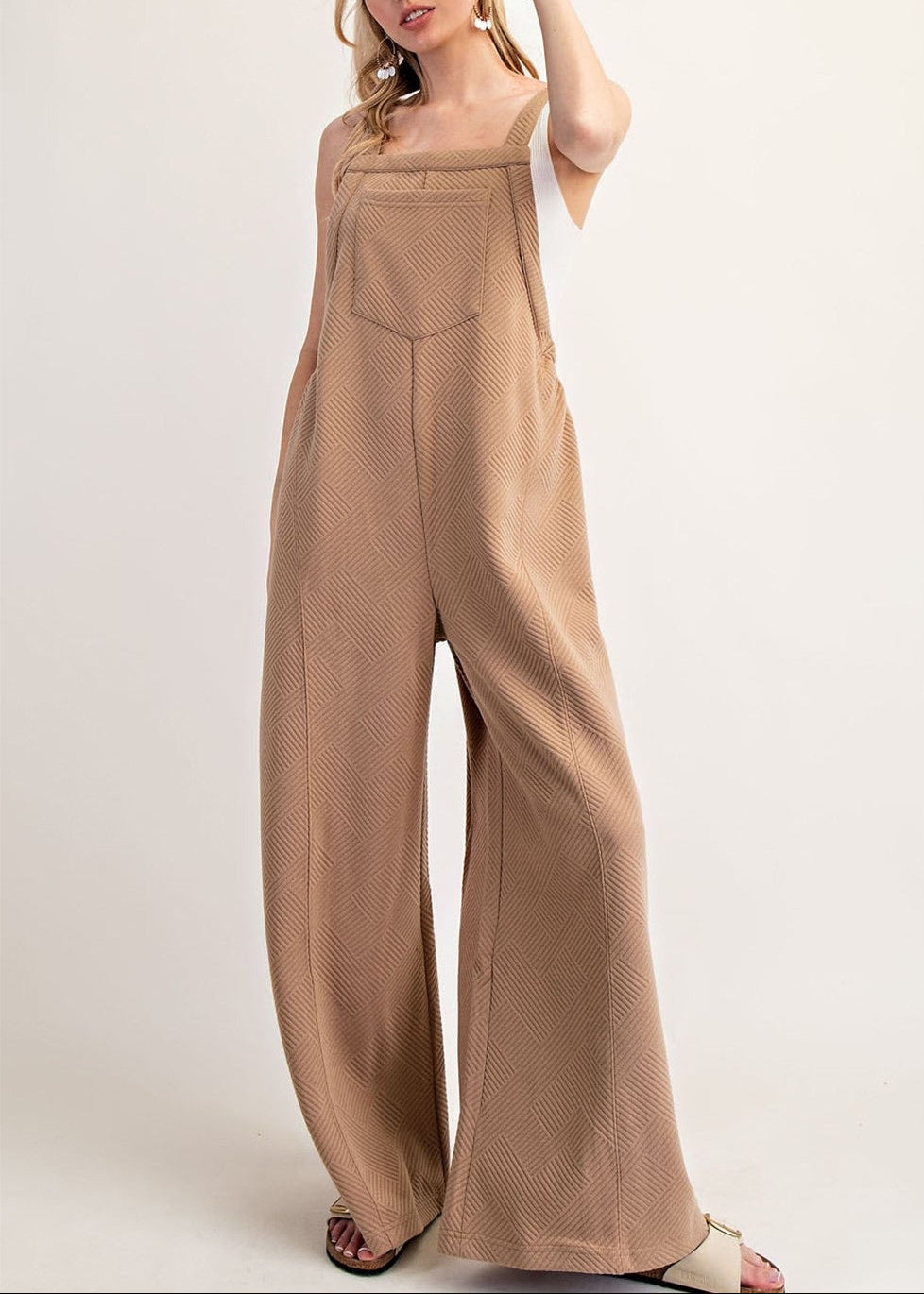 A woman modeling a tan wide-leg jumpsuit with a textured, knit fabric, photographed against a plain off-white studio background.