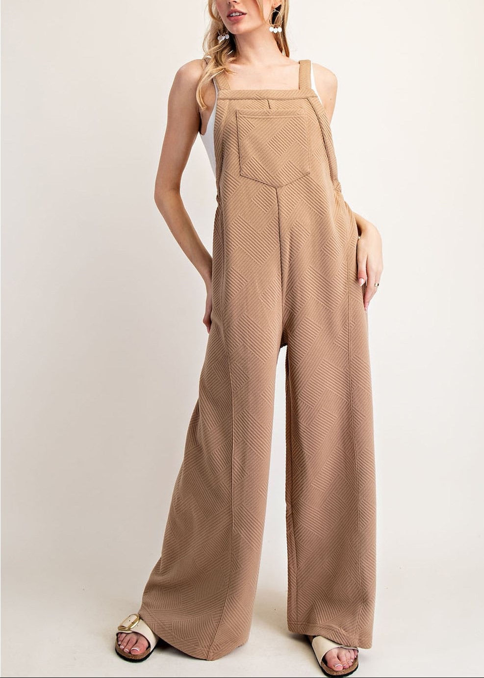 A woman modeling a pair of wide-leg jumpsuit in a textured camel brown fabric, styled with a white tank top and beige sandals against a plain light beige studio background.