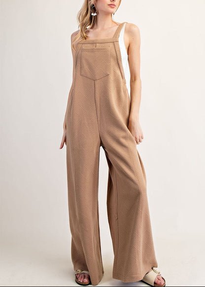 A woman modeling a sleeveless, wide-leg jumpsuit in a warm tan or camel color with a subtle textured pattern, photographed against a plain light beige studio background.