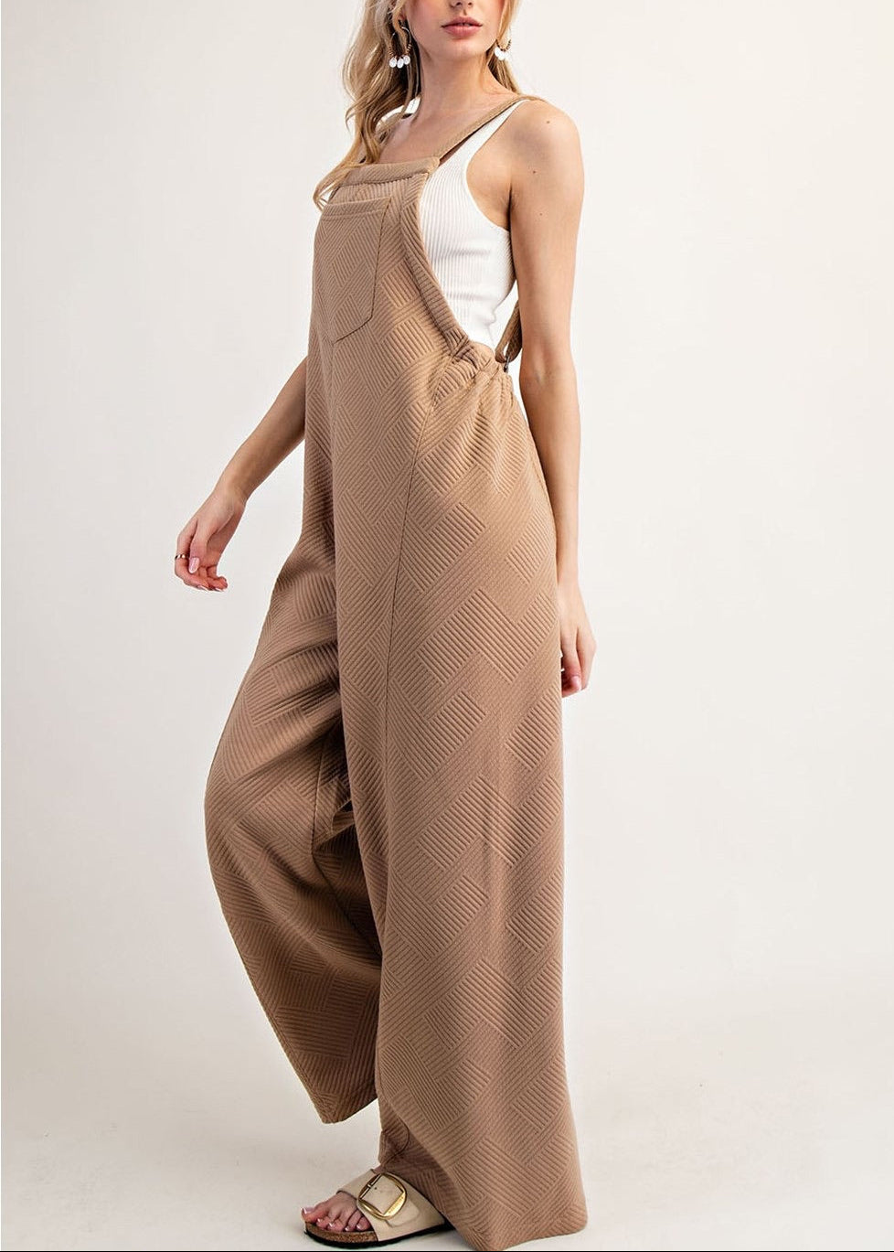 Side view of a woman modeling a pair of wide-leg brown jumpsuit-style overalls with a textured pattern, worn over a white camisole top, against a plain light background.
