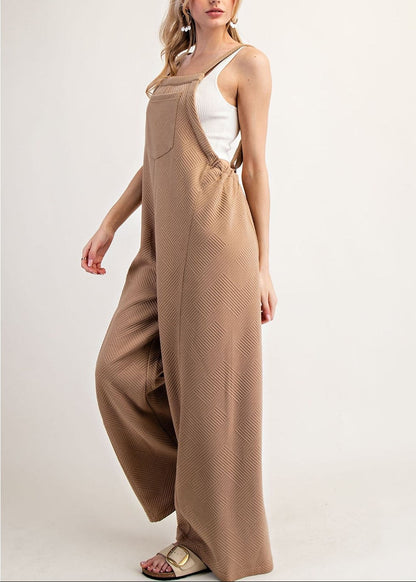 Side view of a woman modeling a pair of wide-leg brown jumpsuit-style overalls with a textured pattern, worn over a white camisole top, against a plain light background.