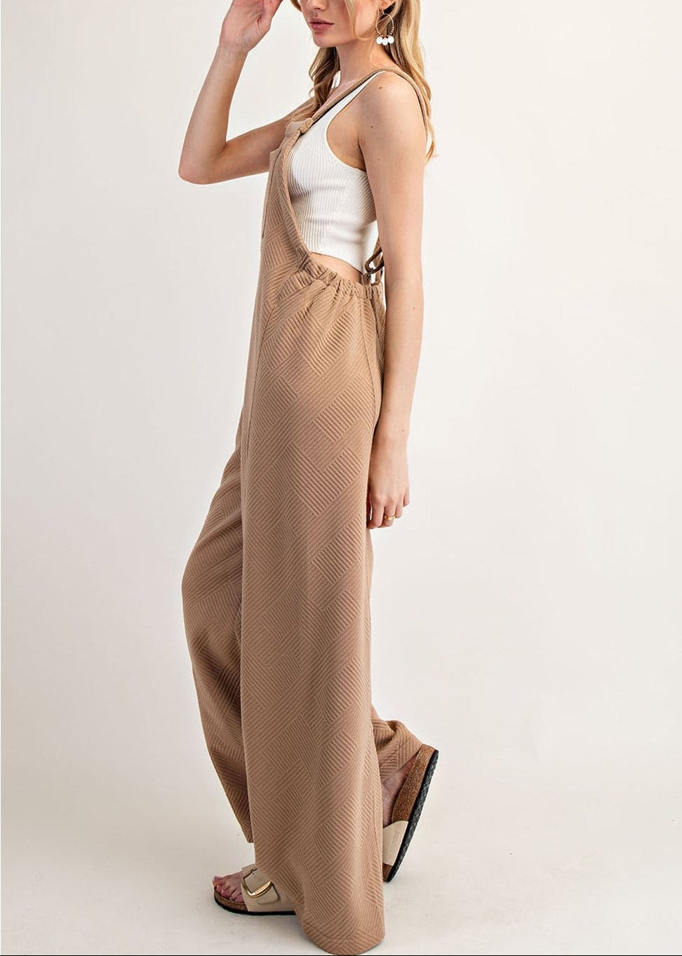 Side view of a woman modeling a pair of wide-leg tan jumpsuit overalls with a white tank top underneath, standing against a plain off-white studio background.