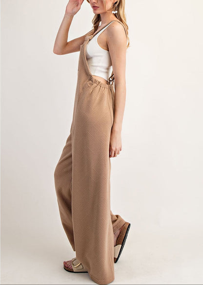 Side view of a woman modeling a pair of wide-leg tan jumpsuit overalls with a white tank top underneath, standing against a plain off-white studio background.