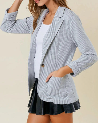 Woman wearing a grey textured blazer over a white top and black skirt on a beige background.