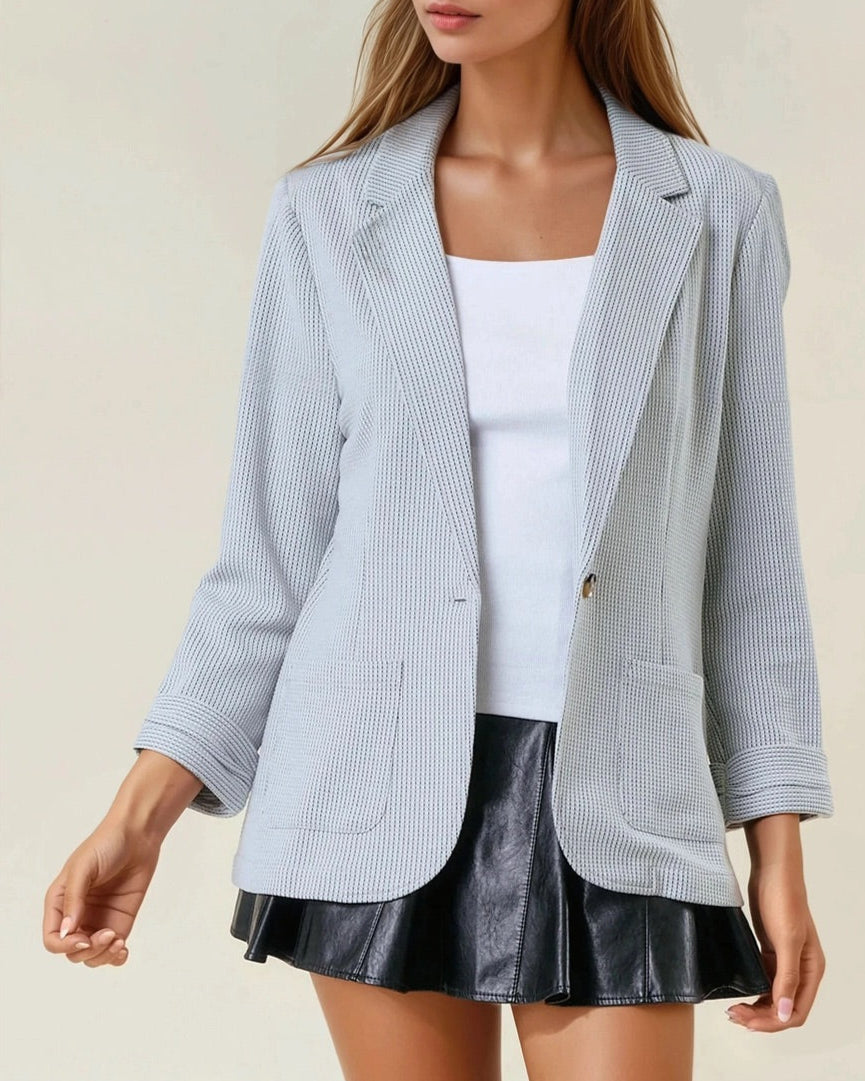 Woman wearing a grey textured blazer over a white top and black skirt on a beige background.