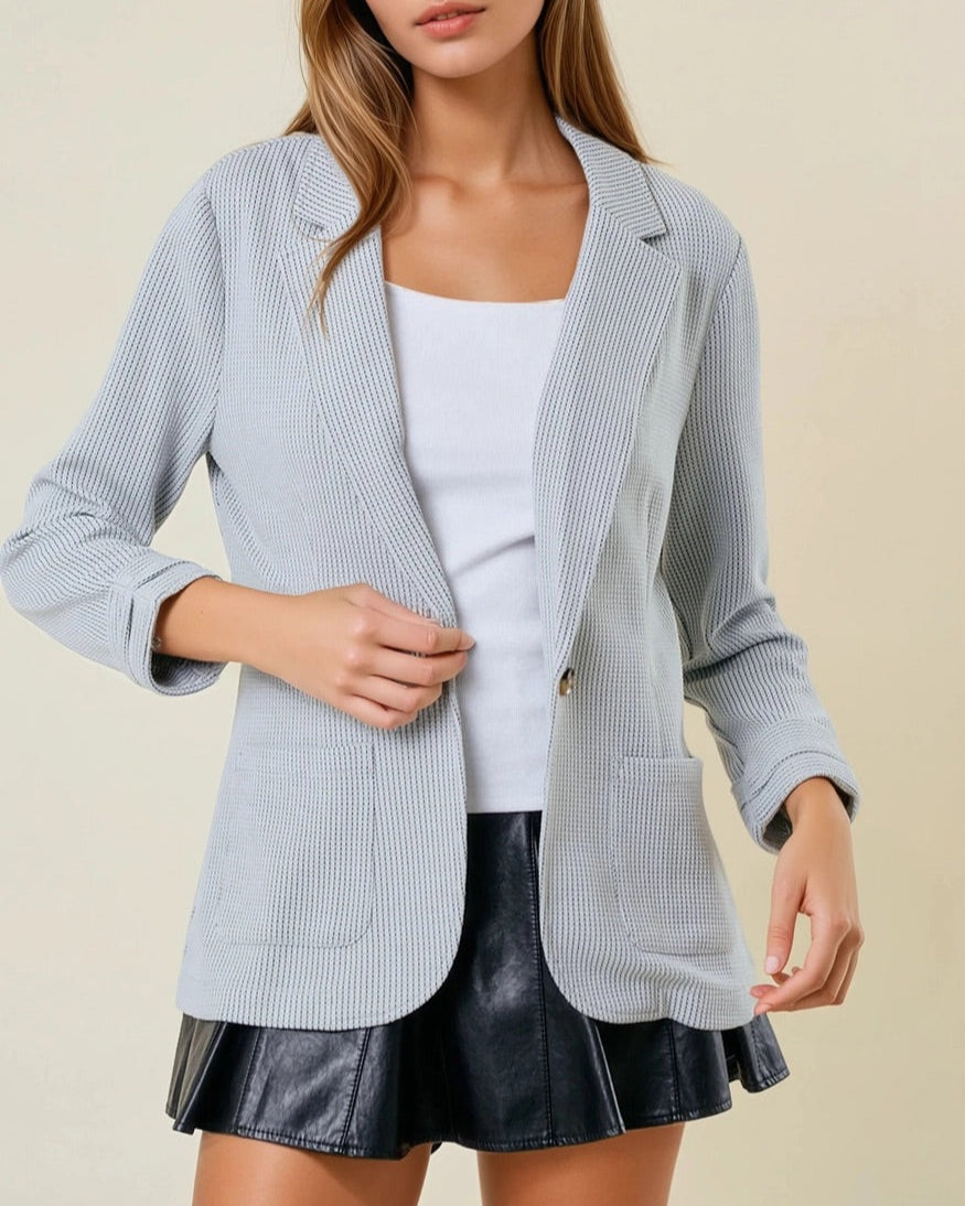 Woman wearing a grey textured blazer over a white top and black skirt on a beige background.