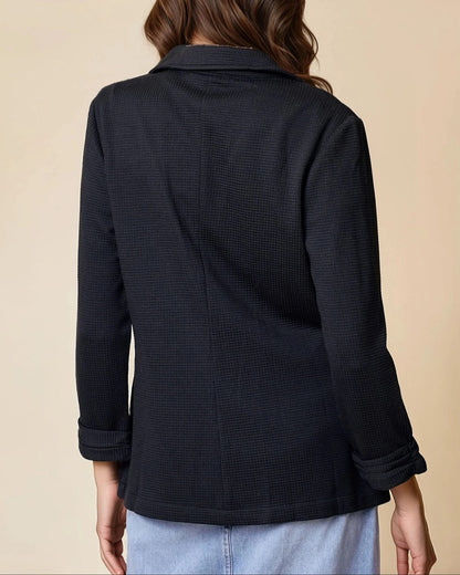 Woman wearing a black textured blazer over a white top and jean skirt on a beige background. 