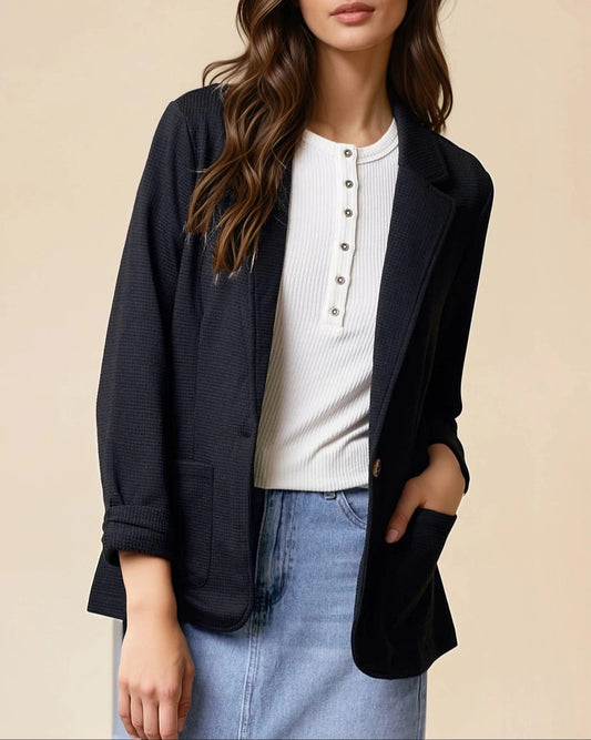 Woman wearing a black textured blazer over a white top and jean skirt on a beige background.