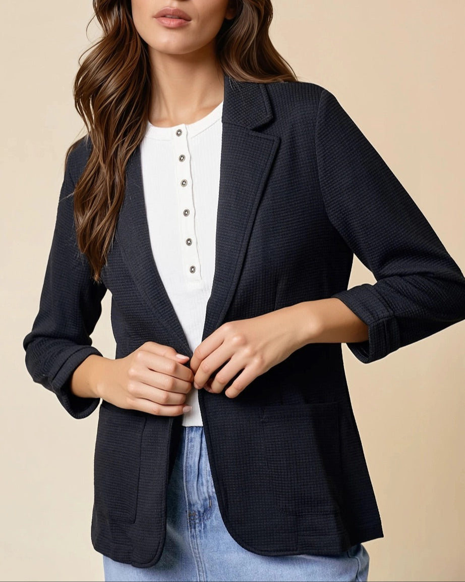 Woman wearing a black textured blazer over a white top and jean skirt on a beige background.
