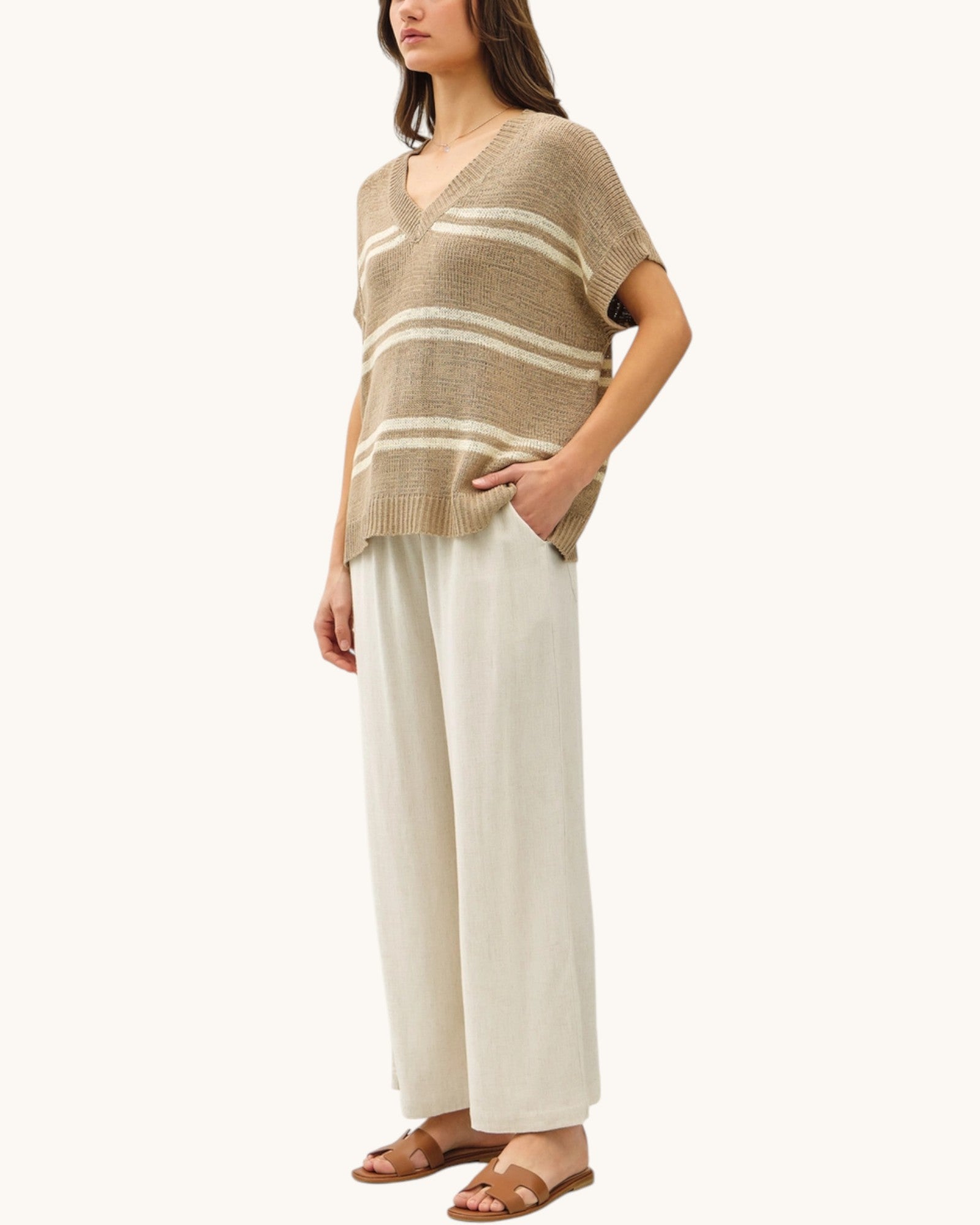 Woman wearing a taupe and white striped short sleeve sweater with wide-leg pants and brown sandals on a white background.