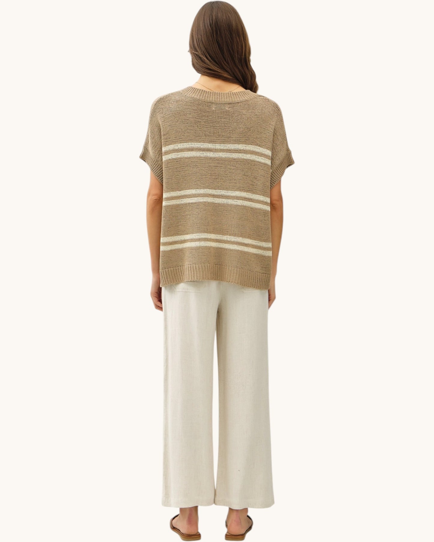Back view of a woman wearing a taupe and cream striped sweater with white pants on a white background.