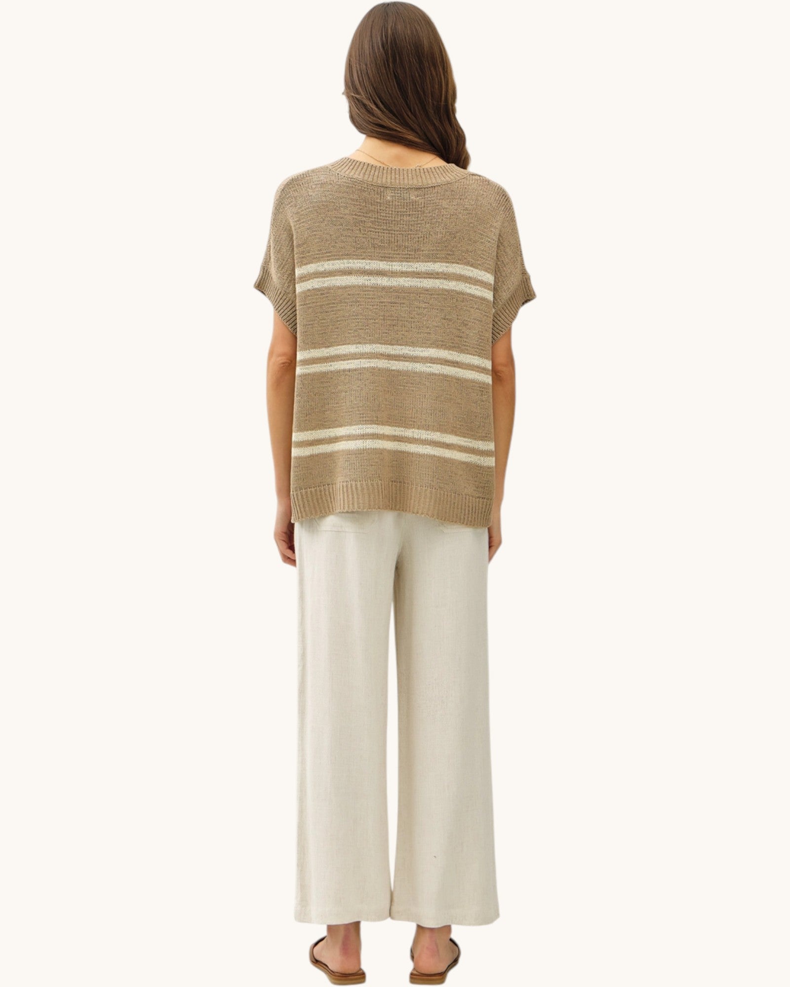 Back view of a woman wearing a taupe and cream striped sweater with white pants on a white background.