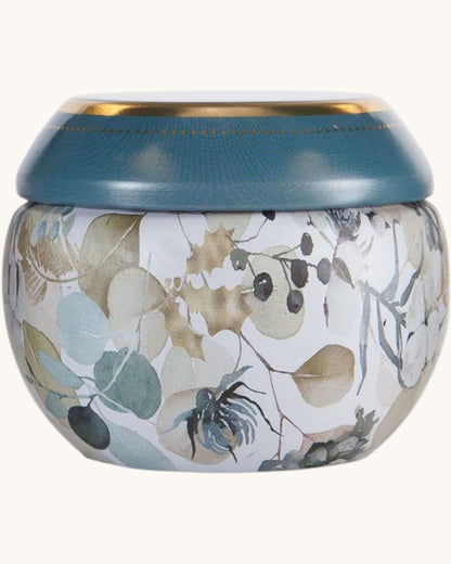 A round decorative jar with a textured teal-blue lid and a white body decorated with a botanical pattern of leaves and berries in muted beige, gray, and blue-green tones.