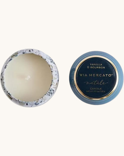 An open round candle tin showing cream-colored wax inside, alongside its matching blue and gold lid printed with 'VIA MERCATO natale' and 'VANILLA & BOURBON'.