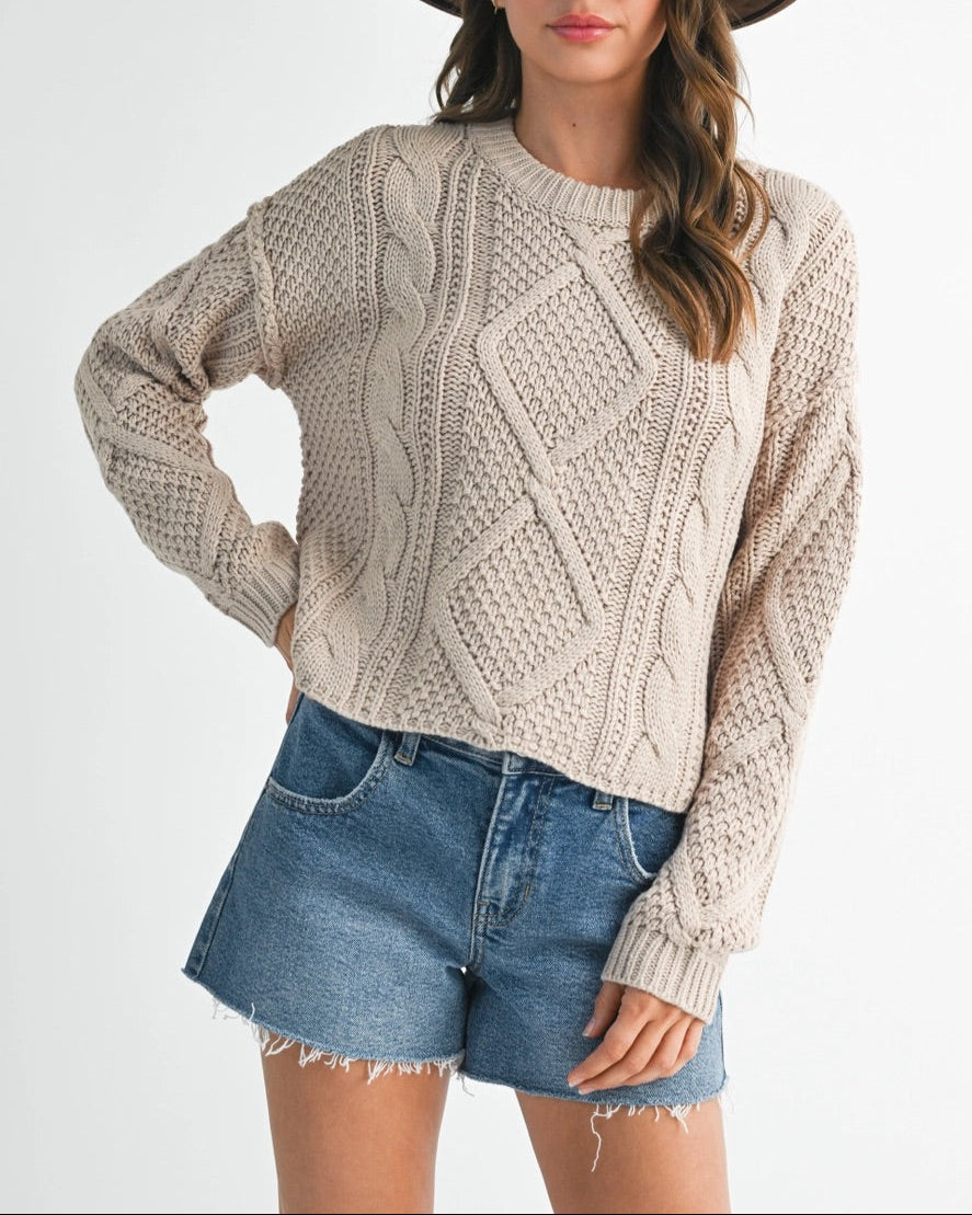 Woman wearing a beige cable knit sweater, denim shorts, and a brown hat on a white background.