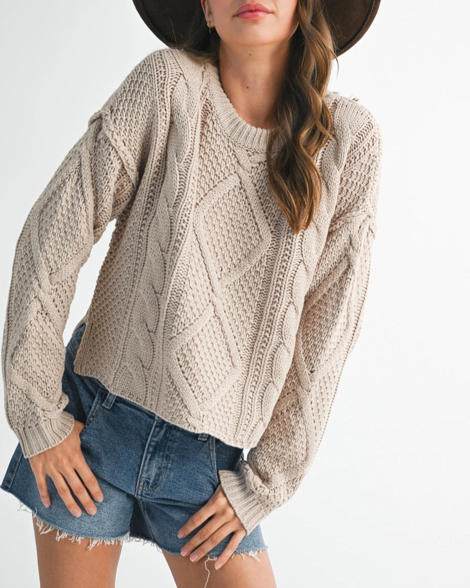 Woman wearing a beige cable knit sweater and denim shorts with a brown hat on a white background