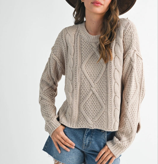 Woman wearing a beige cable knit sweater, denim shorts, and a brown hat on a white background.