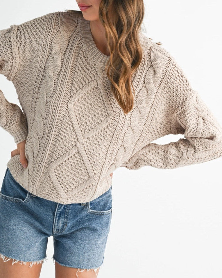 Woman wearing a beige cable knit sweater and denim shorts with a brown hat on a white background.