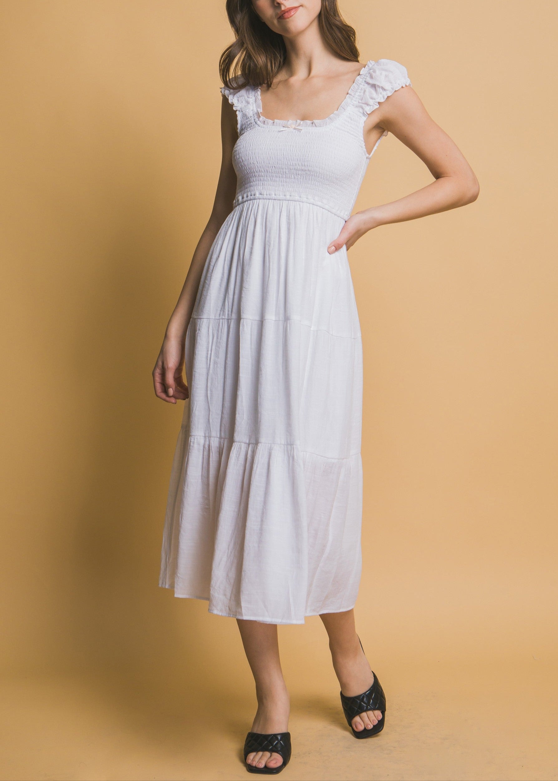 White tiered midi dress with smocked bodice, ruffle straps, and a small rosette ribbon detail at the neckline.