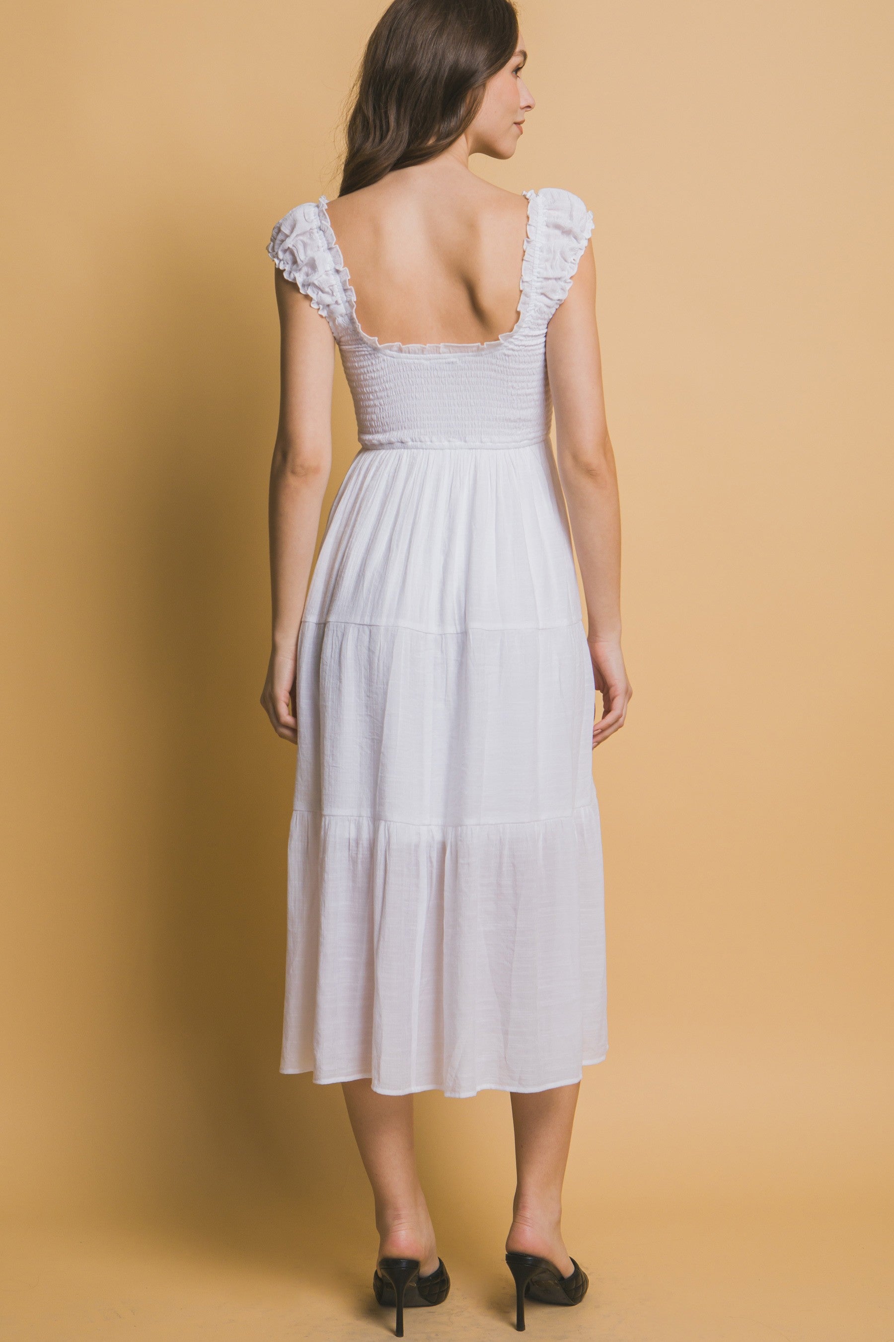 White tiered midi dress with smocked bodice, ruffle straps, and a small rosette ribbon detail at the neckline.