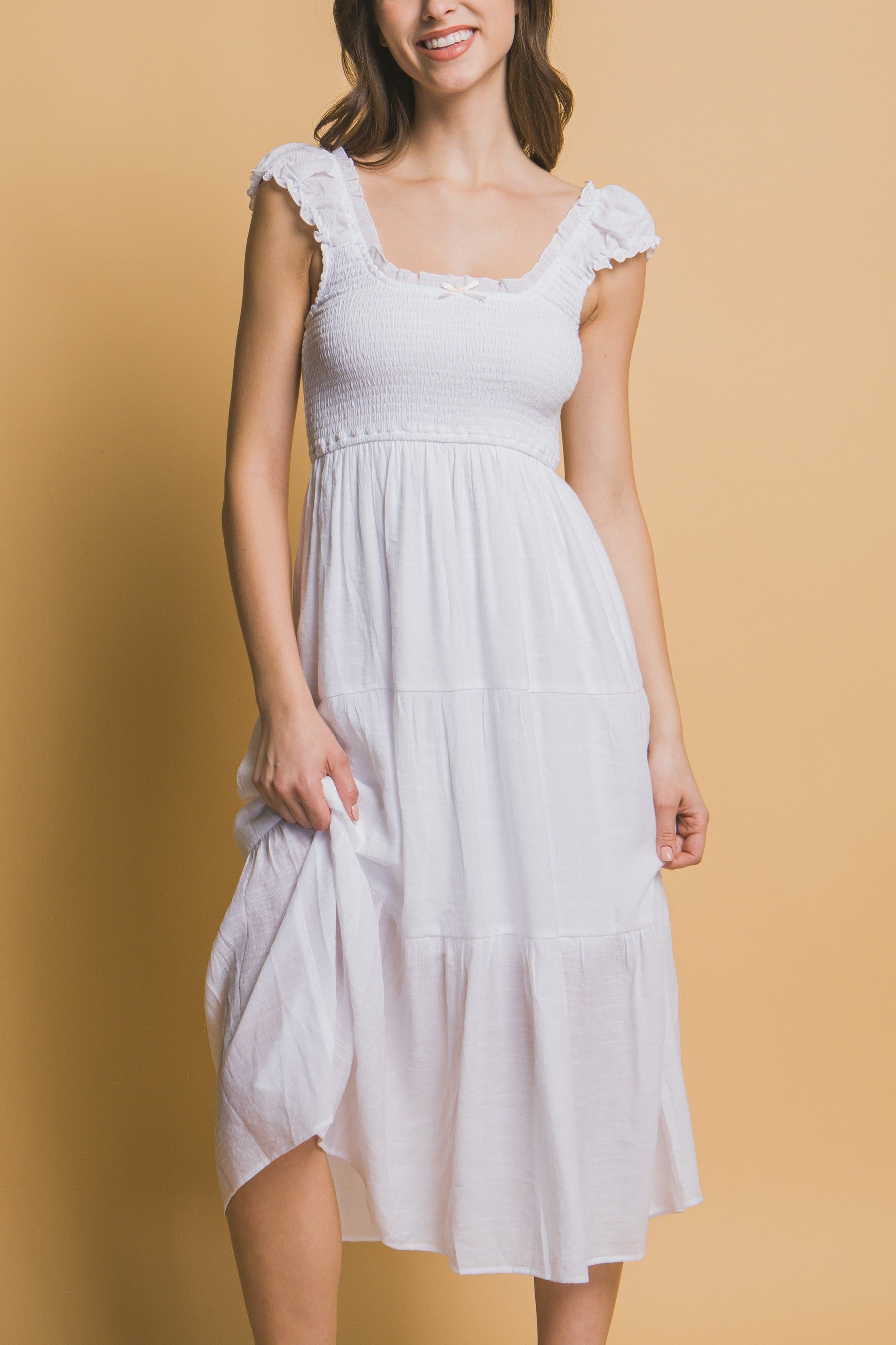 White tiered midi dress with smocked bodice, ruffle straps, and a small rosette ribbon detail at the neckline.