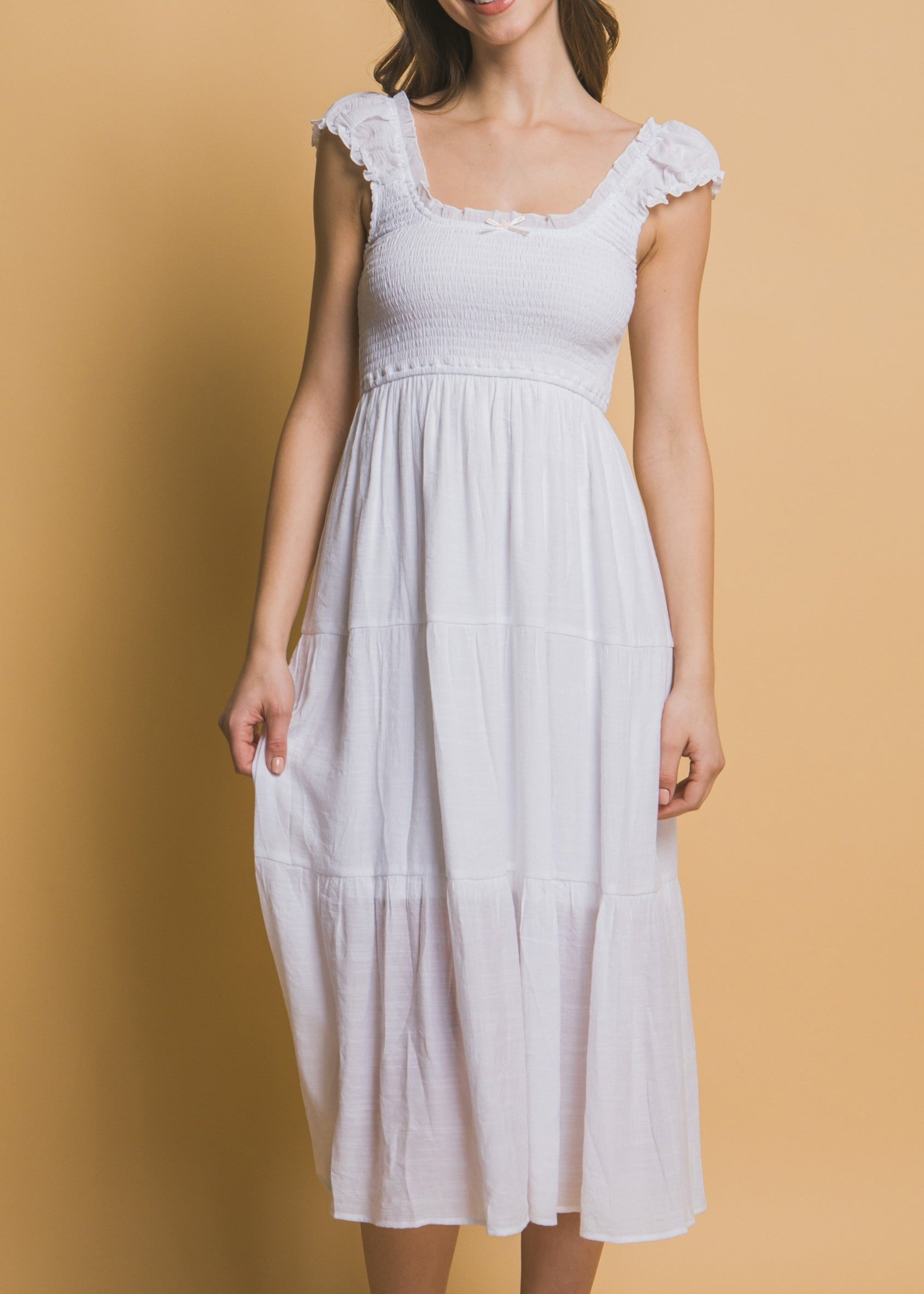 White tiered midi dress with smocked bodice, ruffle straps, and a small rosette ribbon detail at the neckline.