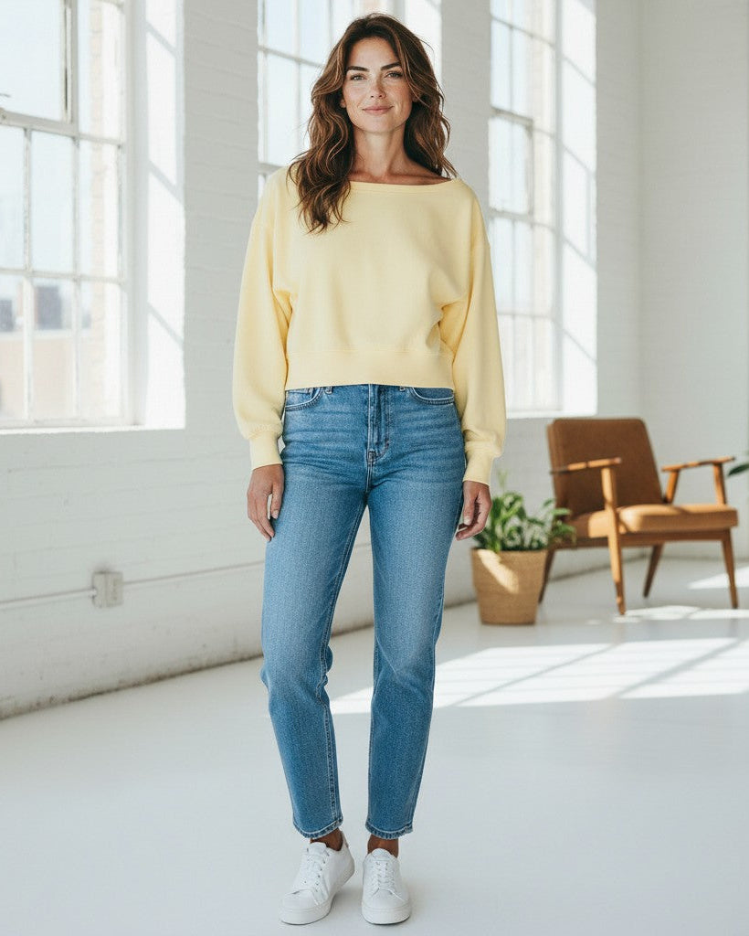 Woman wearing a yellow sweatshirt and blue jeans standing in a bright room with large windows.