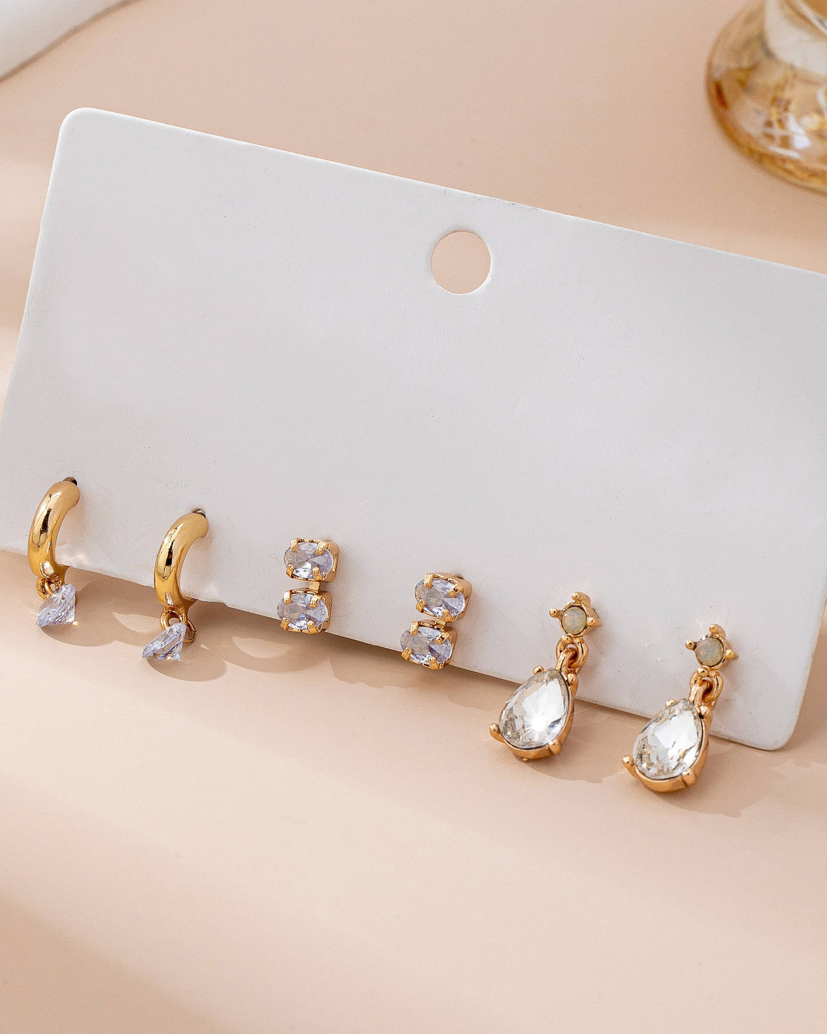 Set of gold and crystal earrings on a white card with a beige background.